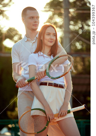 Beautiful young redhead man and woman in stylish, casual, light clothes posing with tennis racket on warm day during sunset time Beautiful young redhead man and woman in stylish, casual, light clothes posing with tennis racket on warm day during sunset time 104399367