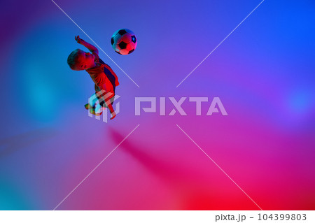 Aerial view of playing little boy during training with football ball in gradient red-blue neon light. Concept of action, sportive lifestyle, team game, health, energy Aerial view of playing little boy during training with football ball in gradient red-blue neon light. Concept of action, sportive lifestyle, team game, health, energy 104399803