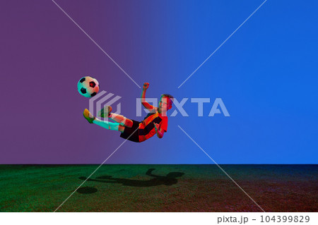 Studio shot of little boy, soccer player in sports uniform jumping with football ball over dark blue background in neon. Concept of sport, health, action, ad Studio shot of little boy, soccer player in sports uniform jumping with football ball over dark blue background in neon. Concept of sport, health, action, ad 104399829