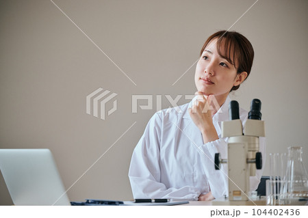 デスクで顎に手を当てて考え事をする白衣の若い女性 104402436