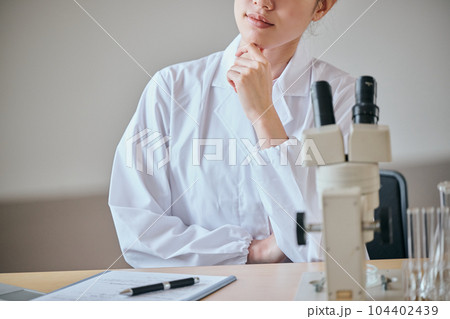 デスクで顎に手を当てて考え事をする白衣の若い女性 104402439
