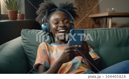 African millennial woman in sweater wearing wireless headphones, listening music, holding and using smart phone, resting and lying on couch in living room. Girl spend lazy weekend home. Generative AI 104405828