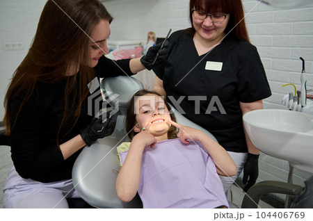 Adorable kid patient, lovely girl pointing her forefingers at her beautiful smile while a dental appointment in pediatric dentistry clinic. Female dentist doctor, dental hygienist examining baby teeth 104406769