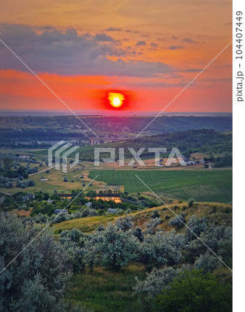 Red summer sunset scenic view over the green valley with bushes and trees. Evening natural landscape Red summer sunset scenic view over the green valley with bushes and trees. Evening natural landscape 104407449