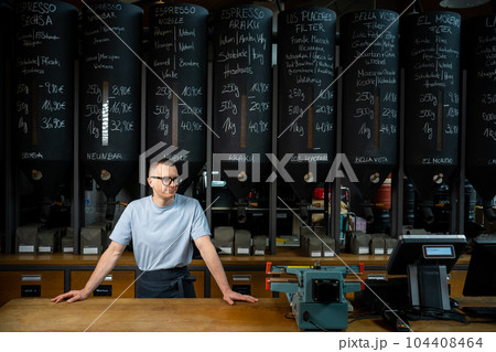 Coffeemaker wearing apron and glasses standing near counter of his cafe with big variety of coffee. Coffeemaker wearing apron and glasses standing near counter of his cafe with big variety of coffee. 104408464