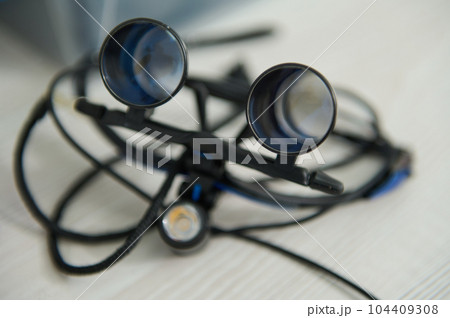 Orthodontists glasses with binocular lenses in dentistry. Dentist eyeglasses, dental optic. Spectacles magnifying binocular for maxillofacial surgery. Modern technology equipment in dental practice Orthodontists glasses with binocular lenses in dentistry. Dentist eyeglasses, dental optic. Spectacles magnifying binocular for maxillofacial surgery. Modern technology equipment in dental practice 104409308