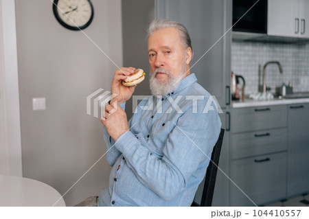 Portrait of handsome bearded mature elderly male eating unhealthy fast food enjoying dinner at home looking at camera. Hungry senior man eating alone delicious burger sitting at table in kitchen room. Portrait of handsome bearded mature elderly male eating unhealthy fast food enjoying dinner at home looking at camera. Hungry senior man eating alone delicious burger sitting at table in kitchen room. 104410557