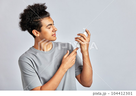 Young handsome smiling guy with black curly hair holding a bottle of facial mask cream in his hands. Man in studio caring for the skin to maintain his youthful and healthy appearance. Men's self-care. Young handsome smiling guy with black curly hair holding a bottle of facial mask cream in his hands. Man in studio caring for the skin to maintain his youthful and healthy appearance. Men's self-care. 104412301