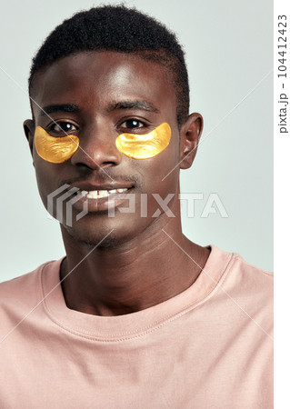 African millennial guy practicing skincare and grooming with under eye hydrogel patches in a studio. Young black man smiling, absorbing the ingredients of the cosmetic under eye mask. Men's self-care. African millennial guy practicing skincare and grooming with under eye hydrogel patches in a studio. Young black man smiling, absorbing the ingredients of the cosmetic under eye mask. Men's self-care. 104412423