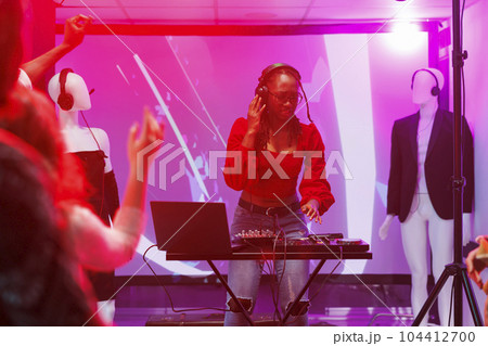Woman dj mixing electronic music using digital controller in nightclub. African american musician in headphones performing on stage with sound mixer console at club party Woman dj mixing electronic music using digital controller in nightclub. African american musician in headphones performing on stage with sound mixer console at club party 104412700