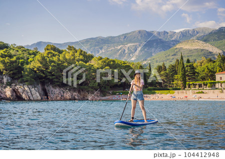 Young women Having Fun Stand Up Paddling in blue water sea near st stefan island against the backdrop of Milocer Park in Montenegro. SUP Young women Having Fun Stand Up Paddling in blue water sea near st stefan island against the backdrop of Milocer Park in Montenegro. SUP 104412948
