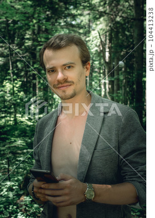 Black and white portrait of Self assured smiling young handsome man with naked chest and open grey jacket. Macho with dark hair, sideburns and beard with phone in park, looking at camera. Black and white portrait of Self assured smiling young handsome man with naked chest and open grey jacket. Macho with dark hair, sideburns and beard with phone in park, looking at camera. 104413238