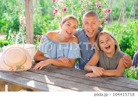 Happy family sits at table in backyard of summer cottage Happy family sits at table in backyard of summer cottage 104415718