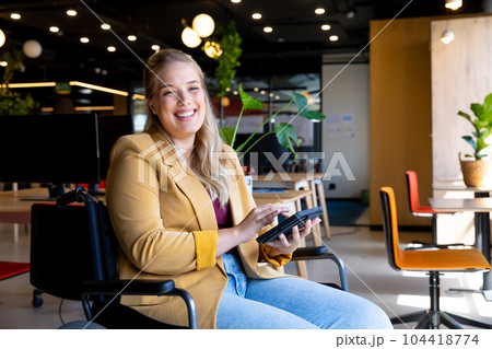 Portrait of happy plus size caucasian casual businesswoman in wheelchair using tablet in office 104418774