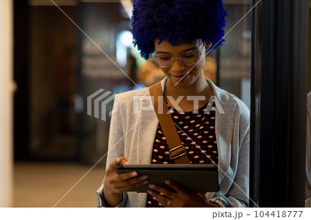 Happy biracial casual businesswoman with blue afro using tablet in office corridor Happy biracial casual businesswoman with blue afro using tablet in office corridor 104418777
