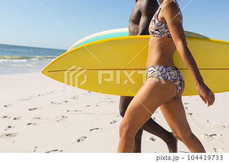 Midsection of fit african american couple carrying surfboards walking on sunny beach to sea Midsection of fit african american couple carrying surfboards walking on sunny beach to sea 104419733