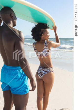 Happy african american couple holding surfboard on heads, smiling on sunny beach by sea Happy african american couple holding surfboard on heads, smiling on sunny beach by sea 104419779