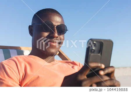 Happy african american man in sunglasses sitting in deckchair using smartphone on sunny beach Happy african american man in sunglasses sitting in deckchair using smartphone on sunny beach 104419888