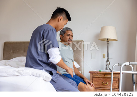 Diverse male doctor and senior male patient sitting on bed and discussing at home 104420225