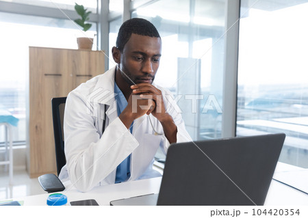 African american male doctor wearing lab coat and stethoscope, using laptop at hospital African american male doctor wearing lab coat and stethoscope, using laptop at hospital 104420354