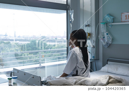 African american female patient sitting on bed and looking through window in hospital room African american female patient sitting on bed and looking through window in hospital room 104420365