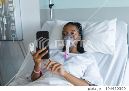 African american female patient with oxygen mask, lying on bed and using smartphone in hospital room 104420390