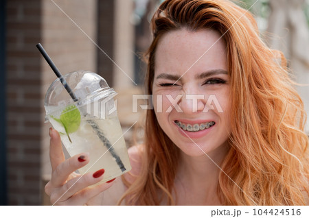 Young beautiful red-haired woman with braces drinks cooling lemonade outdoors in summer. Portrait of a smiling girl with freckles. 104424516
