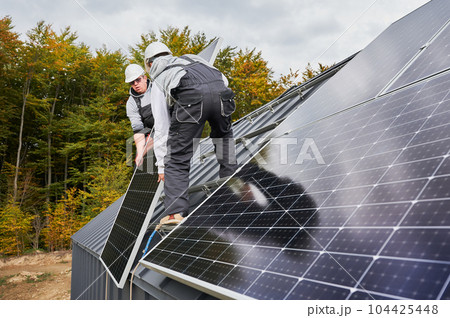 Men roofers installing solar panel system on roof of house. Technicians in helmets lifting up photovoltaic solar module with help of ropes outdoors. Concept of alternative and renewable energy. Men roofers installing solar panel system on roof of house. Technicians in helmets lifting up photovoltaic solar module with help of ropes outdoors. Concept of alternative and renewable energy. 104425448