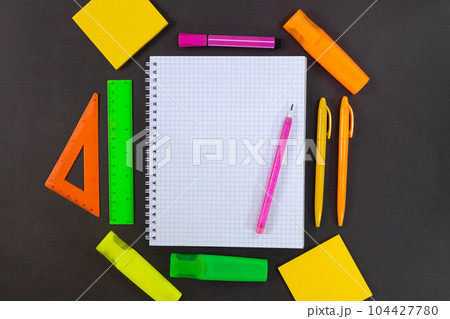 Back to school. On black table lies notebook checkered with pencil for text with empty space. All around are green, yellow, pink, orange stationery. Rulers, markers, felt tip pens, pens, adhesive note 104427780