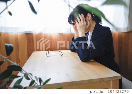 こめかみに手を当てて机に肘をついて座るジャケット姿の女性　疲労　ストレス　悩みを抱えているイメージ 104428270