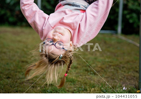 Little preschool girl climbing and hanging on outdoor horizontal bar. Cheerful girl athlete on gymnastic stick on sports ground. Happy child and summer activities on playground. Little preschool girl climbing and hanging on outdoor horizontal bar. Cheerful girl athlete on gymnastic stick on sports ground. Happy child and summer activities on playground. 104431055