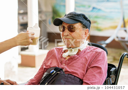 Senior man of 85 years in an electric wheelchair is sitting in a park. Nurse supporting man with drinking coffee, holding cup. Senior man of 85 years in an electric wheelchair is sitting in a park. Nurse supporting man with drinking coffee, holding cup. 104431057