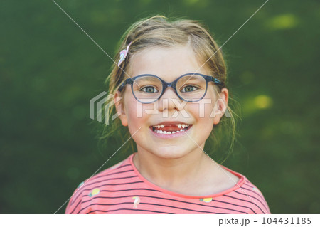 Portrait of a cute preschool girl with eye glasses and big teeth gap outdoors in park. Happy funny child on sunny summer day. Kid loosing milk tooth Portrait of a cute preschool girl with eye glasses and big teeth gap outdoors in park. Happy funny child on sunny summer day. Kid loosing milk tooth 104431185