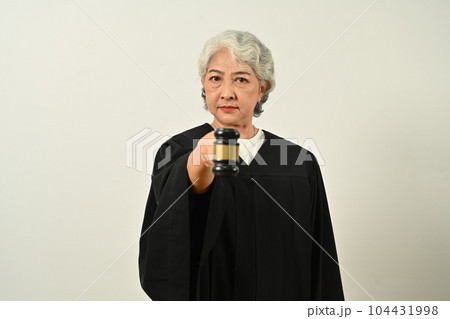 Strict senior female judge dressed in robe gown uniform with gavel hammer mallet isolated on white background. Law and justice concept Strict senior female judge dressed in robe gown uniform with gavel hammer mallet isolated on white background. Law and justice concept 104431998