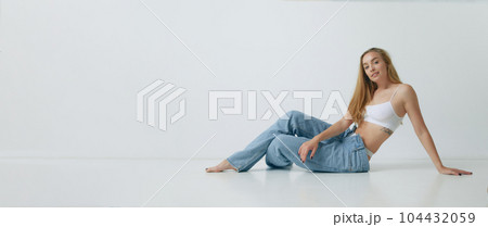 Beautiful young blonde woman with slim body posing in white top and jeans against grey studio background 104432059
