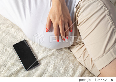 A pregnant girl holds a mobile phone near her belly. The concept of harm and deviations in the development of the unborn child. The negative impact of cellular radiation on pregnant women. 104432110