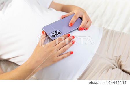 A pregnant girl holds a mobile phone near her belly. The concept of harm and deviations in the development of the unborn child. The negative impact of cellular radiation on pregnant women. 104432111