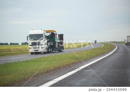White semi-trailer truck trawl transports oversized machinery on the highway in rainy weather, industry. Copy space for text White semi-trailer truck trawl transports oversized machinery on the highway in rainy weather, industry. Copy space for text 104433090