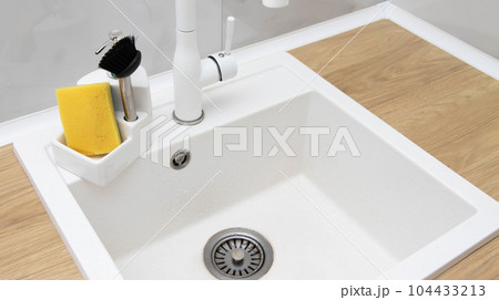 Beautiful kitchen interior of a white sink with a water faucet, sponge and brush for washing dishes and detergent against a white tile wall. Modern interior 104433213