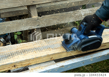 A male carpenter plans boards with an electric planer to make a bench. Carpentry, industry. Woodworker 104433424