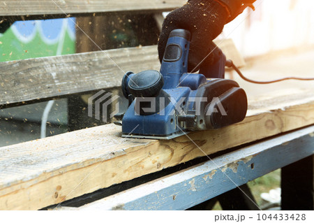 A male carpenter plans boards with an electric planer to make a bench. Carpentry, industry. Woodworker 104433428