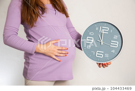 A pregnant girl in the 3rd trimester holds a wall clock against the background of a large belly. The concept of preterm birth, precursors and time to give birth. A pregnant girl in the 3rd trimester holds a wall clock against the background of a large belly. The concept of preterm birth, precursors and time to give birth. 104433469