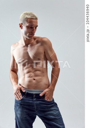 Portrait of handsome young man with blonde hair, musuclar fit body posing shirtless in jeans against grey studio background Portrait of handsome young man with blonde hair, musuclar fit body posing shirtless in jeans against grey studio background 104436940