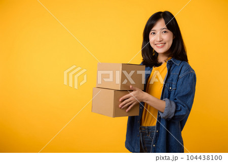 Portrait asian young woman wearing yellow t-shirt and denim shirt holding parcel box isolated on yellow studio background, Delivery courier and shipping service concept. Portrait asian young woman wearing yellow t-shirt and denim shirt holding parcel box isolated on yellow studio background, Delivery courier and shipping service concept. 104438100
