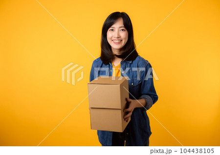 Portrait asian young woman wearing yellow t-shirt and denim shirt holding parcel box isolated on yellow studio background, Delivery courier and shipping service concept. 104438105