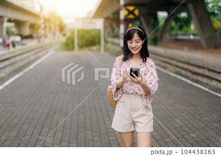 Asian young woman traveler with weaving basket using a mobile phone beside railway train station in Bangkok. Journey trip lifestyle, world travel explorer or Asia summer tourism concept. 104438163
