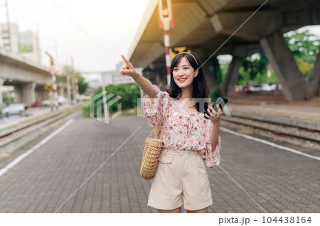 Asian young woman traveler with weaving basket using a mobile phone beside railway train station in Bangkok. Journey trip lifestyle, world travel explorer or Asia summer tourism concept. 104438164