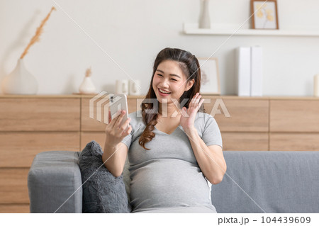 Happy Pregnant Woman sitting on  couch video call conference on mobile phone with family at cozy home.Pregnancy of young woman enjoying with future life relax at home 104439609