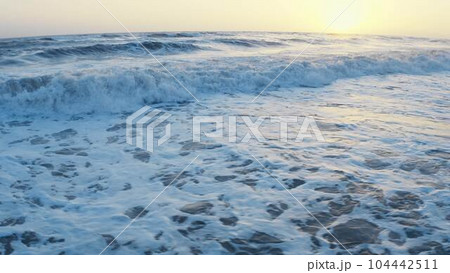 Flying over the ocean during golden hour. Giant waves foaming and splashing in the sea. Cinematic drone shot, sunset wavy seascape. Ocean and sky background  104442511