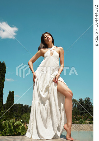 Beautiful brunette woman in a white summer dress on the background of the sky and nature. 104444182